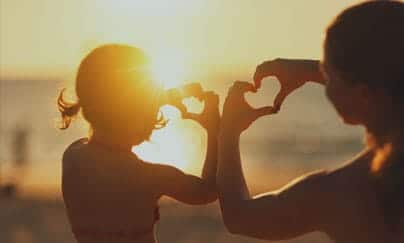 A mother and daughter holding up their hand in shape of a heart against the setting sun over a lake