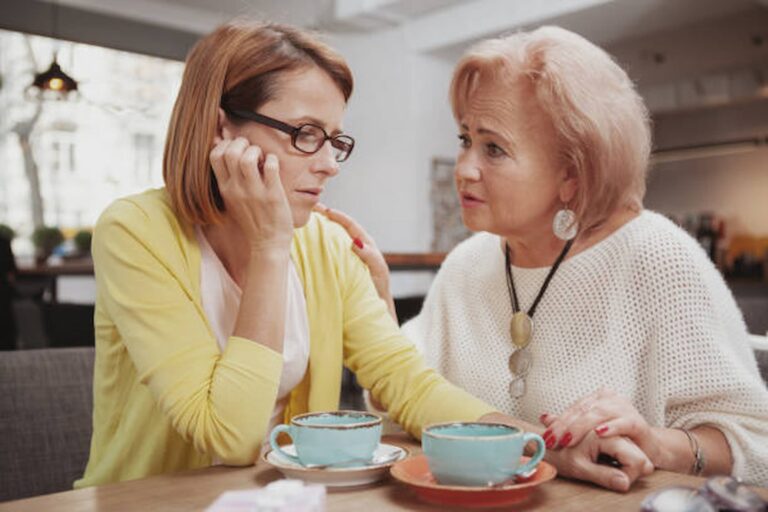 Two women sit at a table with cups of coffee in front of them. In the cozy café in Oregon, one woman, wearing glasses and a yellow cardigan, looks pensive about her recent divorce. The other woman, with short hair and a white top, gently comforts her with a hand on her shoulder.